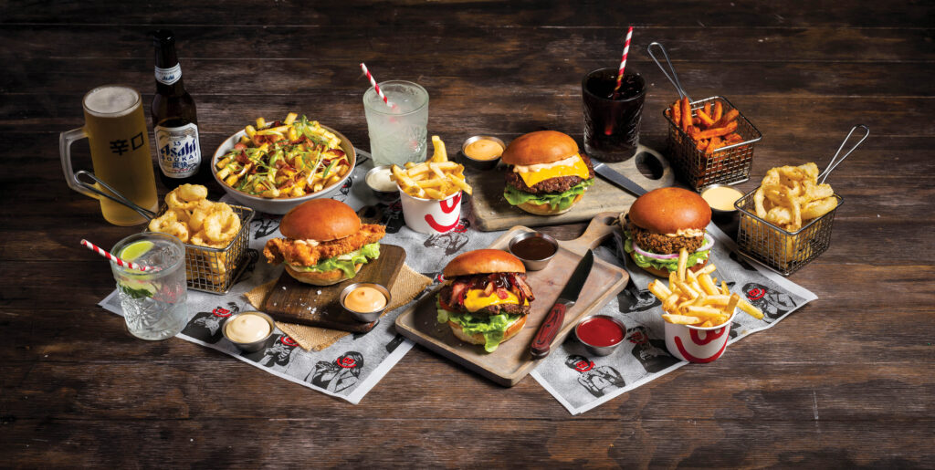 A range of burgers, sides, and drinks spread out over a table at Burger Urge with new paper sitting underneath.