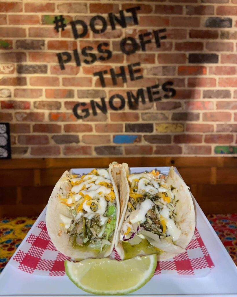 Two tacos sitting on a plate with a decorative wall behind saying 'don't piss off the gnomes' at the Courtyard